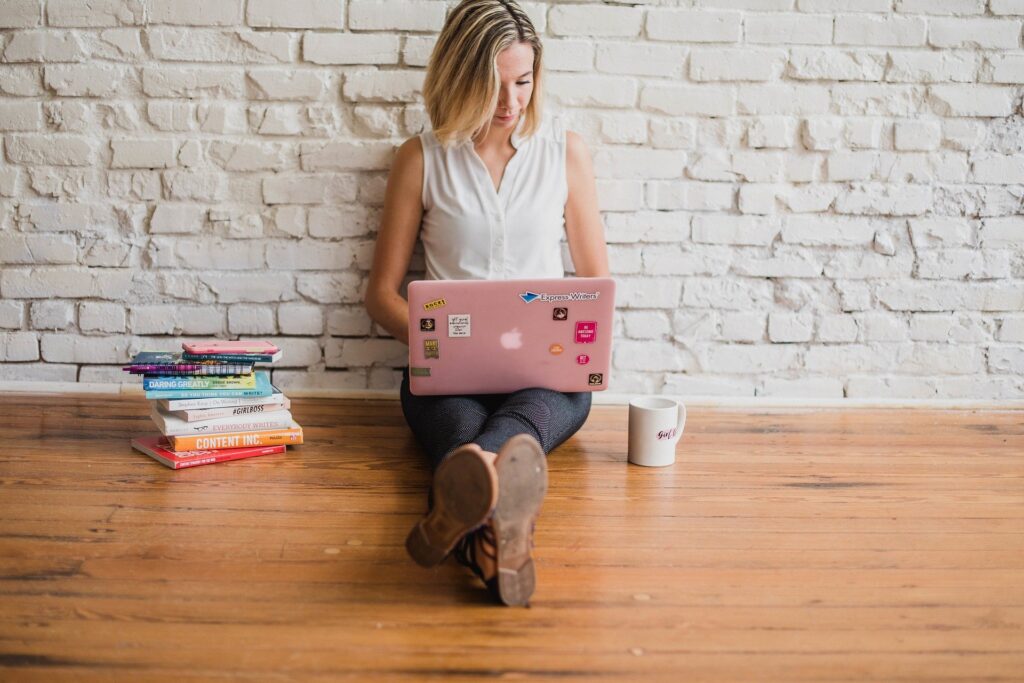 Woman using laptop by books on wooden floor.