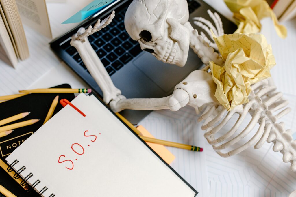 Skeleton passed out on lap top with notes and pencils around it and the message sos.