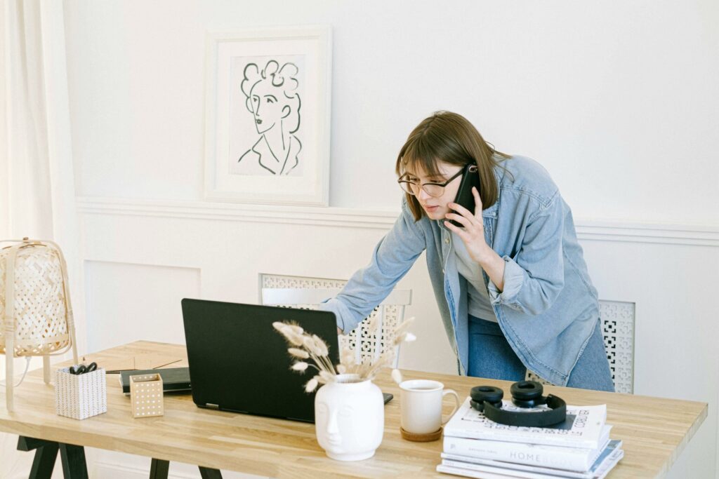 Woman multitasking on laptop and phone in home office.