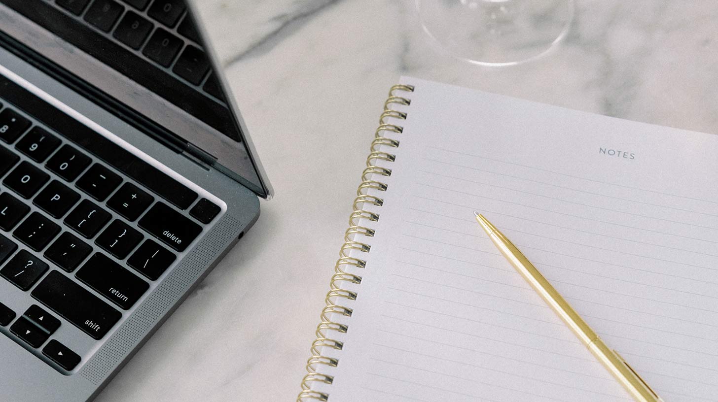 Laptop next to open notebook with pen