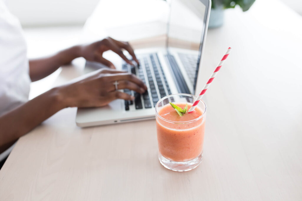 Person typing on laptop with smoothie nearby.