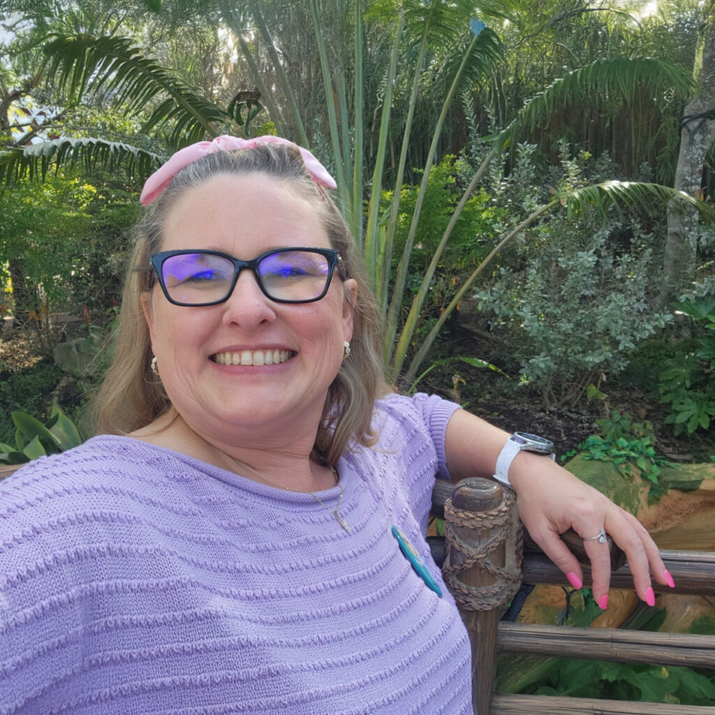 Smiling woman in garden with glasses and purple sweater.