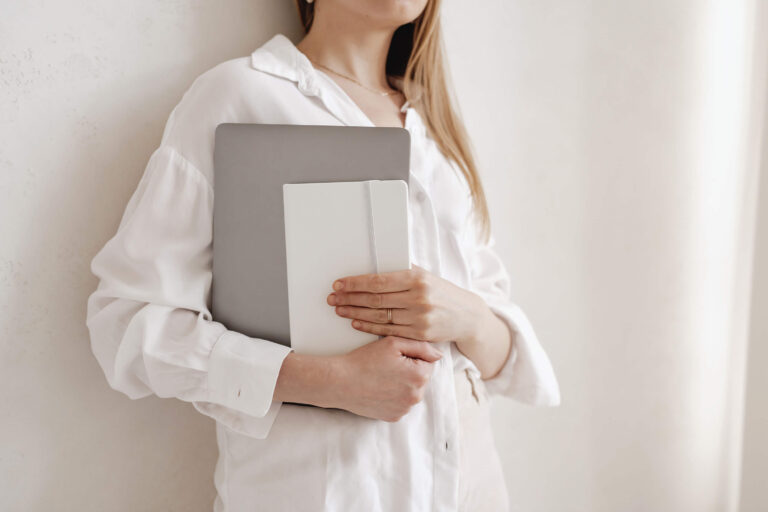 Woman holding laptop and notebook in white blouse.