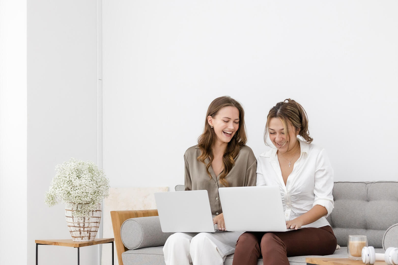 Two women smiling using laptops on a couch.