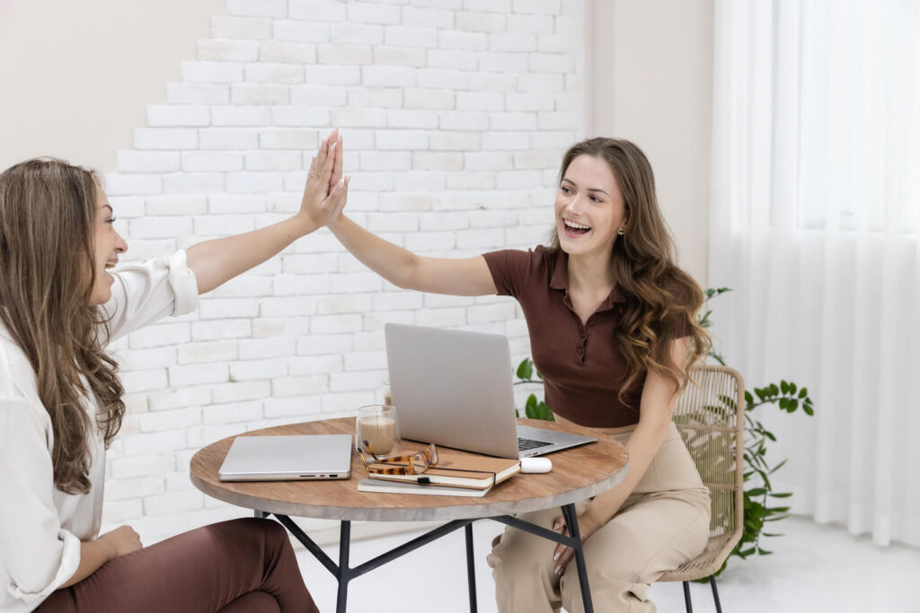 Two women high-fiving at desk with laptops.