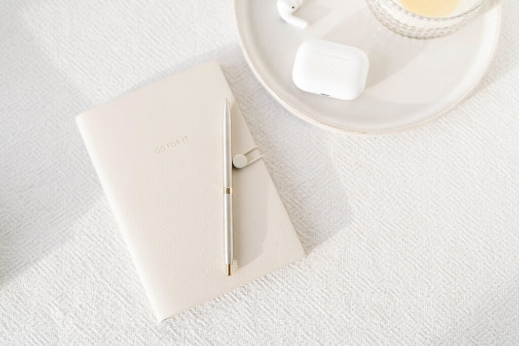 Notebook, pen, AirPods, drink on white cloth surface.