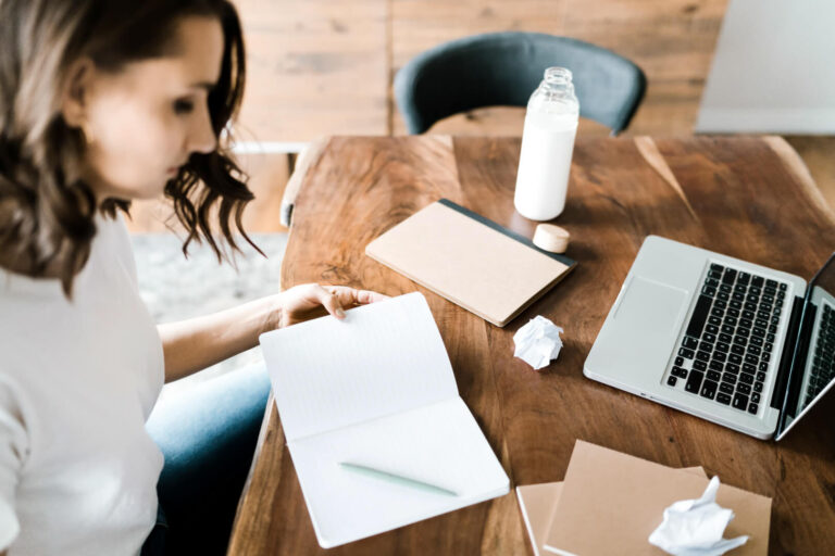 Person writing at desk with laptop and notebook.