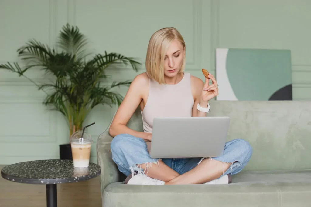 Woman using laptop while eating cookie on couch.