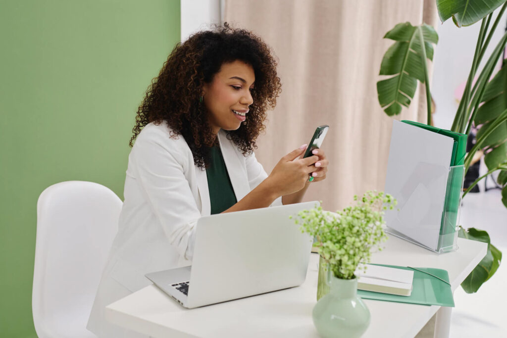 Woman using phone at desk with laptop