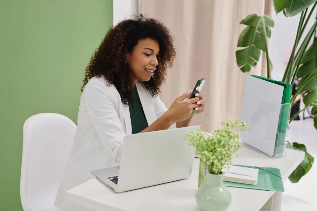Woman using phone at desk with laptop